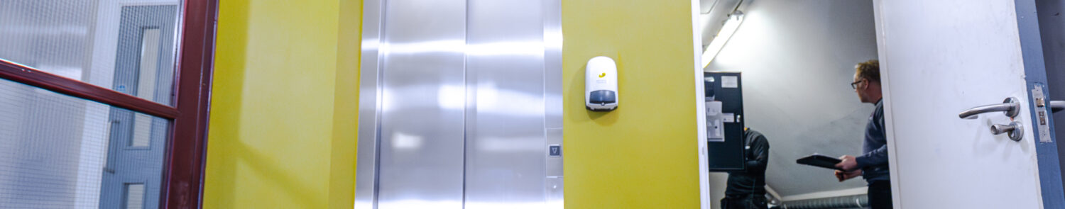 A bright corridor featuring an elevator, a hand sanitiser dispenser, and a person entering a room.