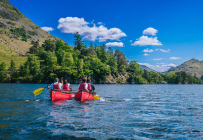 Two people in red kayaks paddle across a calm lake surrounded by lush greenery and blue skies.