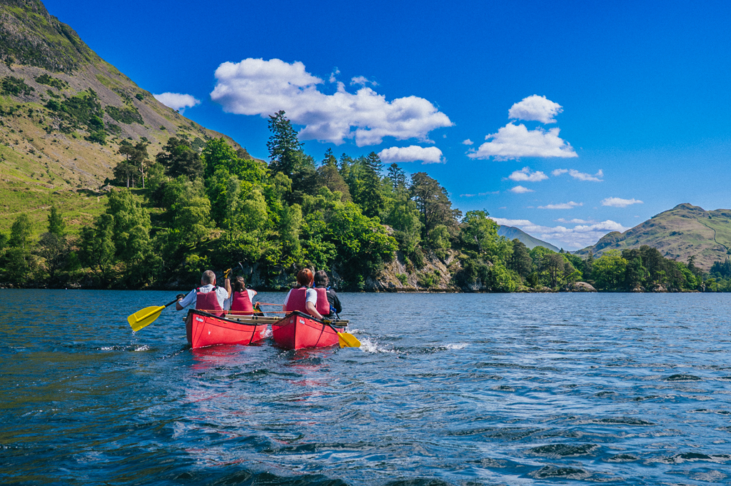 Two people in red kayaks paddle across a calm lake surrounded by lush greenery and blue skies.