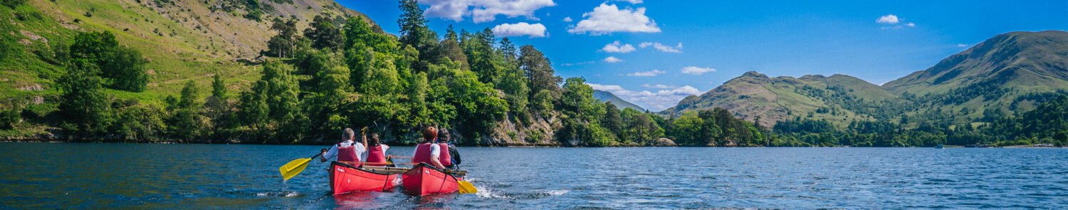 Two people in a red canoe paddle across a calm lake surrounded by lush green hills and blue skies.